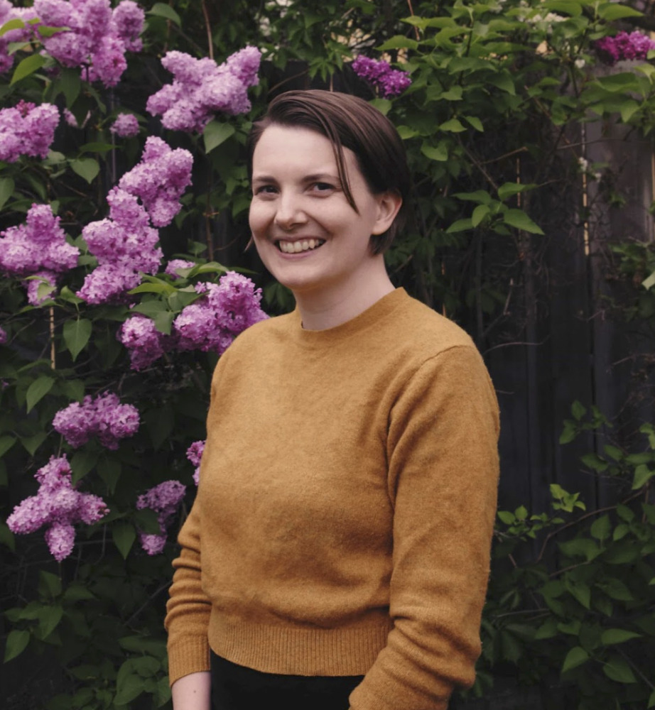 KFPC Food Policy Implementation Lead, Lindsay Harris, wears a yellow sweater in front of a lilac bush.
